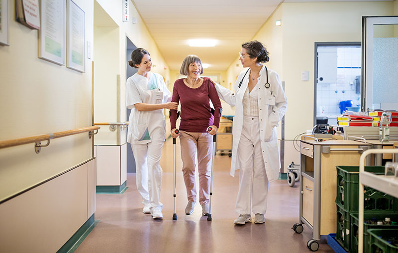 Arzt und Pflegekraft helfen einer älteren Dame freundlich im Krankenhausflur